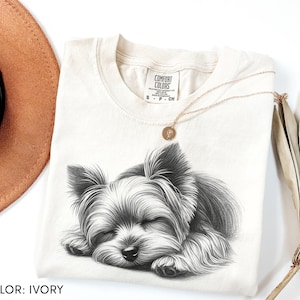 Puede incluir: Camiseta de color marfil con una ilustraci&oacute;n en blanco y negro de un perro durmiendo. La camiseta tiene cuello redondo y mangas cortas. Un collar dorado con un colgante descansa sobre la camiseta. Las palabras "COMFORT COLORS" est&aacute;n impresas en una etiqueta.