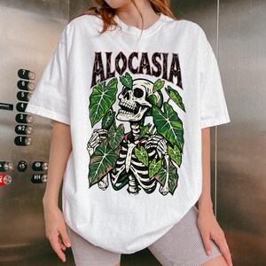 K&ouml;nnte beinhalten: Wei&szlig;es T-Shirt mit dem Wort "ALOCASIA" in schwarzer und roter Schrift. Das Shirt zeigt eine Skelettgrafik, die von gr&uuml;nen Bl&auml;ttern umgeben ist. Das Skelett hat einen detaillierten Sch&auml;del und Brustkorb.