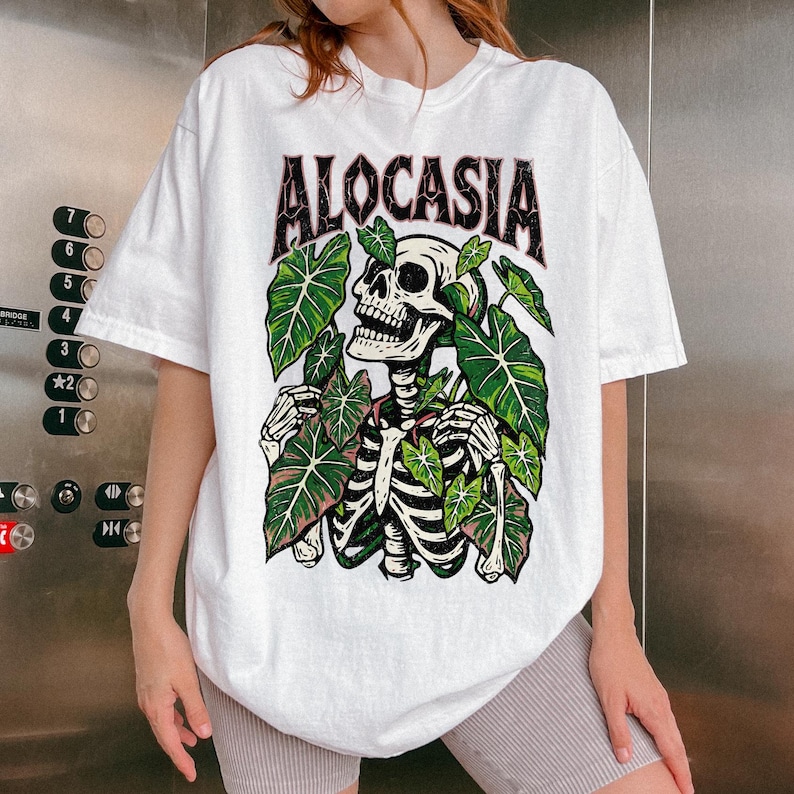 K&ouml;nnte beinhalten: Wei&szlig;es T-Shirt mit dem Wort "ALOCASIA" in schwarzer und roter Schrift. Das Shirt zeigt eine Skelettgrafik, die von gr&uuml;nen Bl&auml;ttern umgeben ist. Das Skelett hat einen detaillierten Sch&auml;del und Brustkorb.