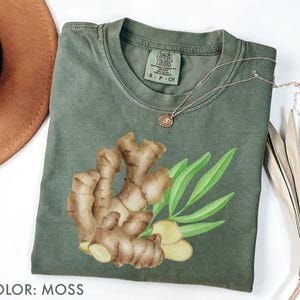 Op de afbeelding: Mosgroen t-shirt met een aquarel illustratie van gemberwortel en bladeren. Het shirt heeft een ronde hals en korte mouwen. Een gouden ketting met een kleine hanger is over het shirt gedrapeerd. De tekst "COLOR: MOSS" staat onderaan.
