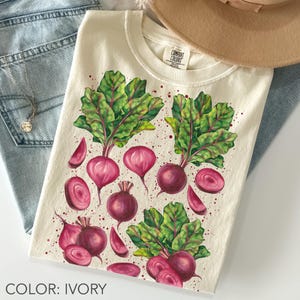 May include: Ivory-colored t-shirt featuring a watercolor illustration of beets with green leaves and red-purple roots. The design includes whole beets and sliced sections. The shirt is folded, with a hat and jeans visible in the background.