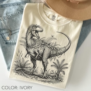 Op de afbeelding: Een ivoorkleurig t-shirt met een gedetailleerde zwarte inktillustratie van een dinosaurus die tussen varens en planten staat. Het shirt heeft een ronde hals en een Comfort Colors merklabel dat aan de binnenkant van de kraag is gedrukt.