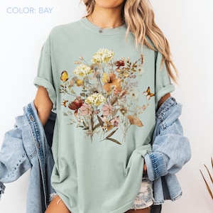 May include: A light green t-shirt featuring a floral design with butterflies. The graphic includes a variety of flowers in shades of yellow, orange, red, and pink. The text "COLOR: BAY" is in the upper left corner.