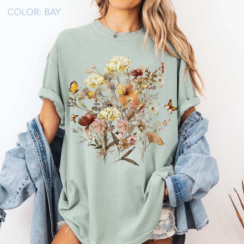 May include: A light green t-shirt featuring a floral design with butterflies. The graphic includes a variety of flowers in shades of yellow, orange, red, and pink. The text "COLOR: BAY" is in the upper left corner.