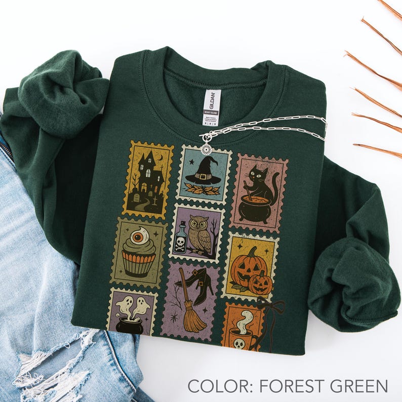 Pu&ograve; includere: Felpa verde foresta con un collage di disegni di francobolli a tema Halloween. I francobolli raffigurano una casa infestata, un cappello da strega, un gatto nero, un cupcake con un bulbo oculare, un gufo, zucche, fantasmi e una scopa da strega. Una collana d'argento &egrave; drappeggiata sulla felpa.