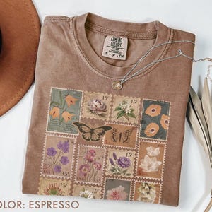 K&ouml;nnte beinhalten: Espressofarbenes T-Shirt mit Rundhalsausschnitt und floralem Stempeldesign. Das Design umfasst Schmetterlinge und verschiedene Blumen in Orange-, Lila- und Rosat&ouml;nen. Eine silberne Halskette mit Anh&auml;nger liegt auf dem T-Shirt.