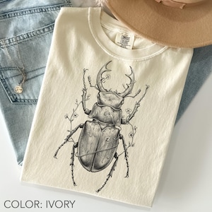 Può includere: T-shirt color avorio con un'illustrazione dettagliata in bianco e nero di un cervo volante e accenti floreali. La maglietta ha un girocollo ed è realizzata in materiale morbido. Il testo "COMFORT COLORS" è visibile sull'etichetta.