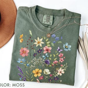 Puede incluir: Una camiseta de color musgo con un dise&ntilde;o floral con flores rosas, amarillas, azules y moradas. La camiseta tiene cuello redondo y mangas cortas. Un collar de oro con un colgante est&aacute; sobre la camiseta. El texto "COLOR: MOSS" est&aacute; en la parte inferior.