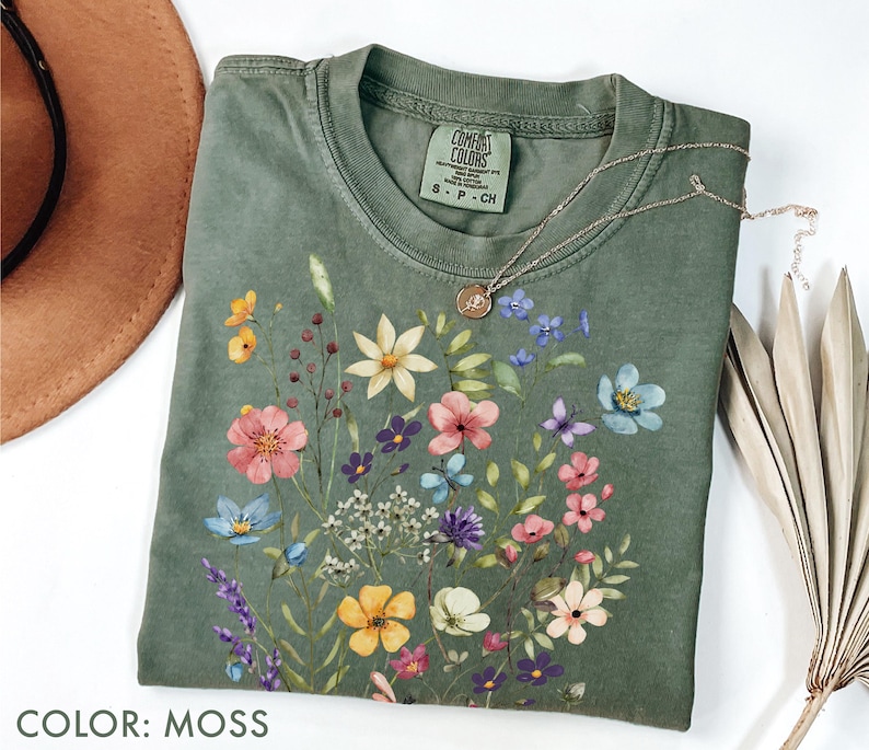 Puede incluir: Una camiseta de color musgo con un dise&ntilde;o floral con flores rosas, amarillas, azules y moradas. La camiseta tiene cuello redondo y mangas cortas. Un collar de oro con un colgante est&aacute; sobre la camiseta. El texto "COLOR: MOSS" est&aacute; en la parte inferior.