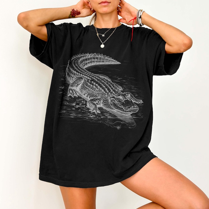 May include: Black t-shirt featuring a detailed grayscale illustration of an alligator. The artwork depicts the alligator in a body of water, with intricate shading and texture. The shirt has a relaxed fit.