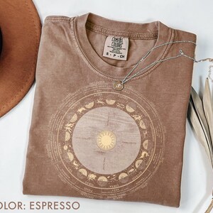Pode incluir: Camiseta cor espresso com um design de zod&iacute;aco dourado. O design apresenta s&iacute;mbolos do zod&iacute;aco e um sol no centro. Um colar de prata com um pequeno pingente est&aacute; sobre a camiseta. A camiseta &eacute; rotulada como "Comfort Colors."