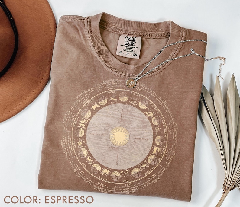 Pode incluir: Camiseta cor espresso com um design de zod&iacute;aco dourado. O design apresenta s&iacute;mbolos do zod&iacute;aco e um sol no centro. Um colar de prata com um pequeno pingente est&aacute; sobre a camiseta. A camiseta &eacute; rotulada como "Comfort Colors."