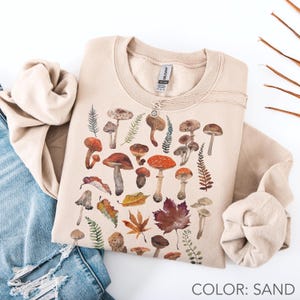Peut inclure: Sweat-shirt couleur sable avec une illustration à l'aquarelle de divers champignons et feuilles d'automne. Le motif comprend des champignons rouges, bruns et beiges, ainsi que des fougères et des feuilles d'érable. Le texte "COLOR: SAND" est visible en bas.