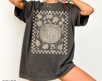 Op de afbeelding: Oversized t-shirt in peperkleur met een beige pompoenillustratie in een geruit kader. Het ontwerp bevat bladeren en kalebassen, wat een herfstthema oproept. Het shirt heeft korte mouwen en een relaxte pasvorm, ideaal voor casual wear.