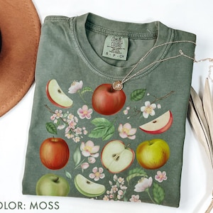 May include: A moss-colored t-shirt featuring a vibrant apple and floral design. The design includes whole and sliced apples in red, green, and yellow, along with delicate pink blossoms and green leaves. A gold necklace rests on the shirt.