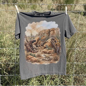 Könnte beinhalten: Ein anthrazitfarbenes Kurzarm-T-Shirt mit einem detaillierten Grafikdruck einer Wüstenlandschaft. Die Illustration zeigt ein Gila-Krustenechse, eine Bartagame und zwei kleinere Eidechsen, die auf felsigem Gelände ruhen.