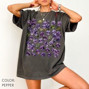 May include: Pepper-colored t-shirt featuring a floral design of purple violets and green leaves. The shirt has a relaxed fit and short sleeves. The text "COLOR: PEPPER" is at the bottom left of the image.