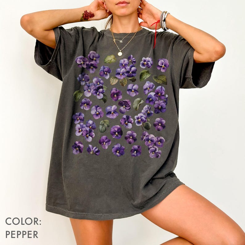 May include: Pepper-colored t-shirt featuring a floral design of purple violets and green leaves. The shirt has a relaxed fit and short sleeves. The text "COLOR: PEPPER" is at the bottom left of the image.