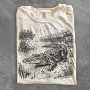 May include: A cream-coloured t-shirt featuring a detailed black ink illustration of an alligator resting on a riverbank near mangrove trees. The graphic includes small crabs, birds in the sky, and the Comfort Colors brand label at the neck.