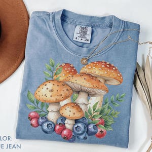 May include: Blue jean coloured t-shirt with a watercolour illustration of mushrooms, blueberries, and leaves. The shirt has a crew neck and short sleeves. A gold necklace with a pendant is draped across the shirt.