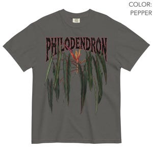 May include: A pepper-colored short-sleeve t-shirt featuring a graphic print of a philodendron plant with long, dark green leaves and a central orange flower. The word PHILODENDRON is printed in a bold, distressed black font above the plant illustration.