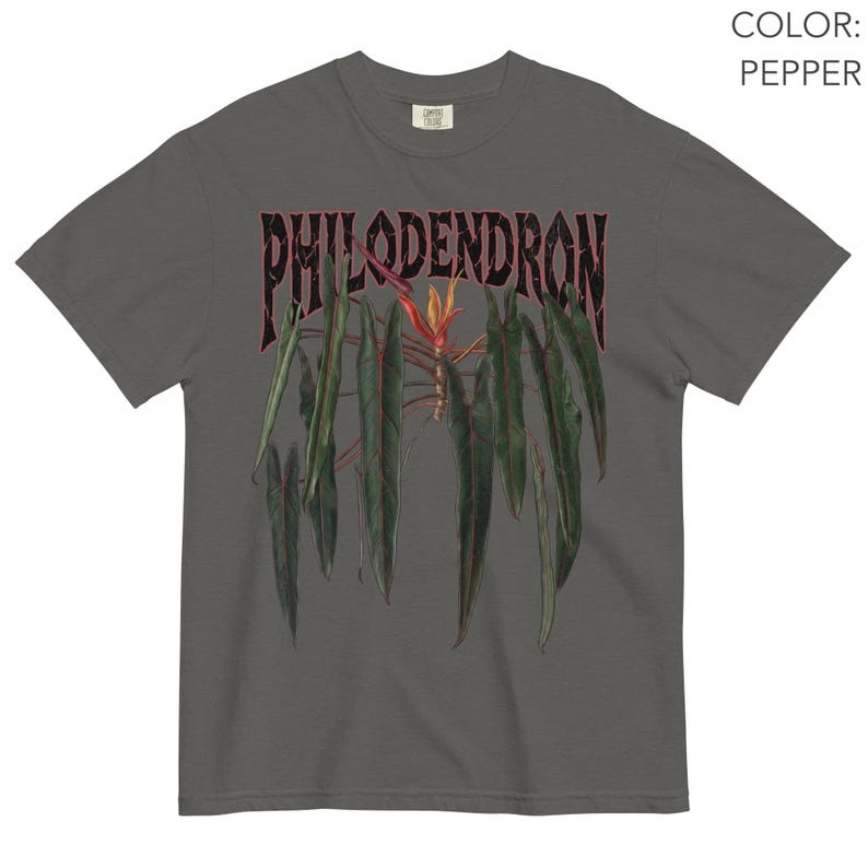 May include: A pepper-colored short-sleeve t-shirt featuring a graphic print of a philodendron plant with long, dark green leaves and a central orange flower. The word PHILODENDRON is printed in a bold, distressed black font above the plant illustration.