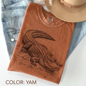 May include: A yam-colored t-shirt featuring a detailed illustration of an alligator. The shirt is paired with light blue jeans and a tan hat. The shirt's design includes the text "XL - TG - EG".