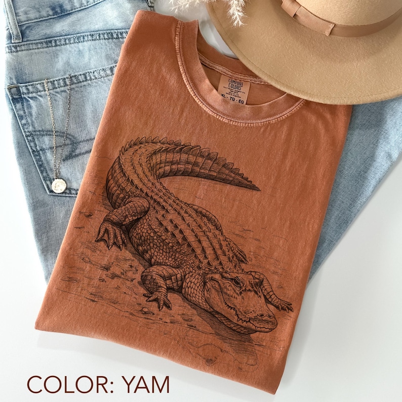 May include: A yam-colored t-shirt featuring a detailed illustration of an alligator. The shirt is paired with light blue jeans and a tan hat. The shirt's design includes the text "XL - TG - EG".