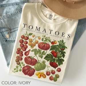 May include: Ivory-colored t-shirt featuring a botanical illustration of various tomato varieties. The design includes red, yellow, and green tomatoes with the text "TOMATOES" and "SOLANUM LYCOPERSICUM - BOTANICAL STUDY". The shirt is paired with blue jeans and a necklace.