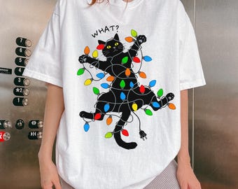 May include: White t-shirt featuring a black cat tangled in colorful Christmas lights. The cat has yellow eyes and the word "WHAT?" is above it. The shirt has a relaxed fit.