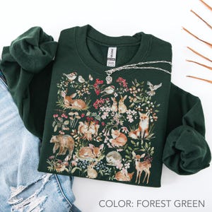 May include: Forest green sweatshirt featuring a detailed woodland scene with various animals, including deer, foxes, and rabbits, surrounded by floral elements. A silver necklace rests on the sweatshirt. The text "COLOR: FOREST GREEN" is at the bottom.