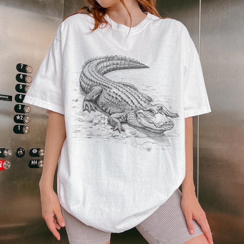 May include: White t-shirt featuring a detailed black and white illustration of an alligator. The alligator is depicted lying down, with its scales and features clearly visible. The shirt has short sleeves and a relaxed fit.