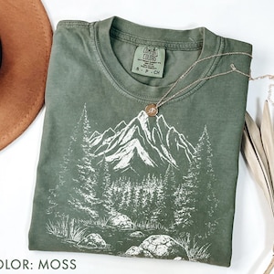 Puede incluir: Una camiseta de color musgo con un gráfico blanco de montaña y bosque. La camiseta tiene cuello redondo y mangas cortas. Un collar dorado con un colgante circular descansa sobre la camiseta. La camiseta es de un material suave y cómodo.