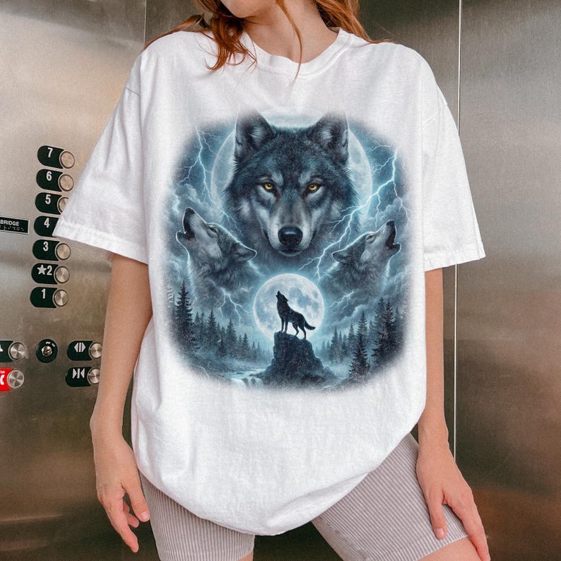 K&ouml;nnte beinhalten: Wei&szlig;es T-Shirt mit einer detaillierten Grafik von W&ouml;lfen, die unter einem Vollmond mit Blitzen heulen. Das Design umfasst ein zentrales Wolfsgesicht und drei heulende W&ouml;lfe, vor dem Hintergrund eines Waldes und eines Nachthimmels.