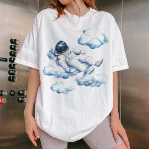 Peut inclure: T-shirt blanc avec une illustration &agrave; l'aquarelle d'un astronaute flottant sur des nuages. L'astronaute porte une combinaison spatiale et un casque, et les nuages sont bleu clair. Le motif est centr&eacute; sur le devant du t-shirt.