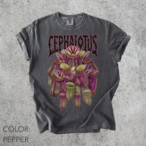 May include: A charcoal gray t-shirt with a graphic print of a cluster of purple and green pitcher plants. Above the plants, the text CEPHALOTUS is printed in a stylized black font. The shirt features a crew neckline and short sleeves.