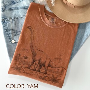 Pu&ograve; includere: Una maglietta color igname con un'illustrazione dettagliata di un dinosauro Brontosauro in un paesaggio preistorico. La maglietta &egrave; una taglia XL-TG-EG Comfort Colors. La maglietta &egrave; abbinata a jeans blu e un cappello marrone.