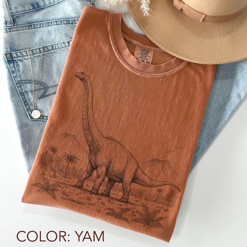 Pu&ograve; includere: Una maglietta color igname con un'illustrazione dettagliata di un dinosauro Brontosauro in un paesaggio preistorico. La maglietta &egrave; una taglia XL-TG-EG Comfort Colors. La maglietta &egrave; abbinata a jeans blu e un cappello marrone.