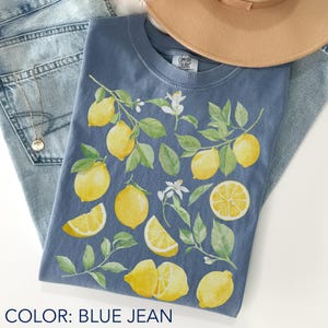 Blue jean Comfort Colors t-shirt featuring watercolor lemons and green leaves in a soft botanical coquette aesthetic, styled flat lay with denim shorts and hat, fresh citrus summer vibe