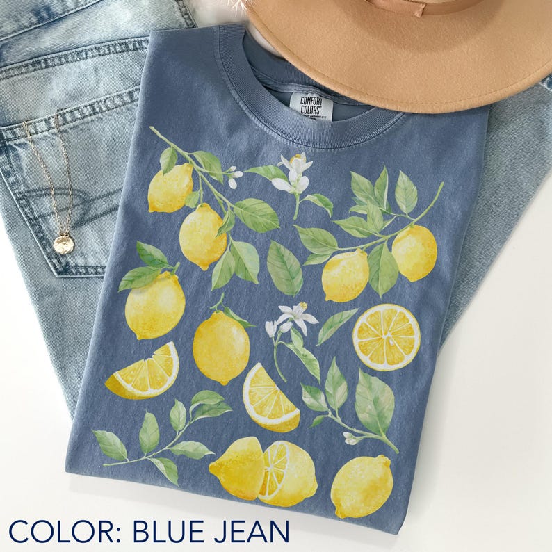 Blue jean Comfort Colors t-shirt featuring watercolor lemons and green leaves in a soft botanical coquette aesthetic, styled flat lay with denim shorts and hat, fresh citrus summer vibe