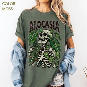K&ouml;nnte beinhalten: Moosgr&uuml;nes T-Shirt mit einem Skelett-Design, umgeben von gr&uuml;nen Bl&auml;ttern. Das Wort "ALOCASIA" ist in Dunkelbraun &uuml;ber dem Skelett aufgedruckt. Das Shirt hat eine lockere Passform.