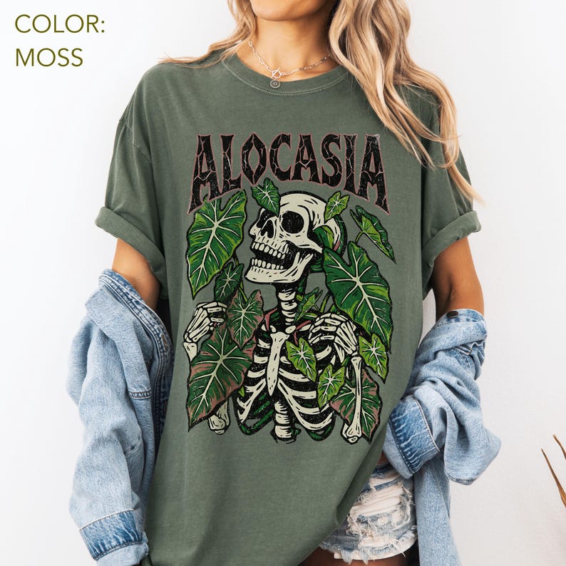 K&ouml;nnte beinhalten: Moosgr&uuml;nes T-Shirt mit einem Skelett-Design, umgeben von gr&uuml;nen Bl&auml;ttern. Das Wort "ALOCASIA" ist in Dunkelbraun &uuml;ber dem Skelett aufgedruckt. Das Shirt hat eine lockere Passform.