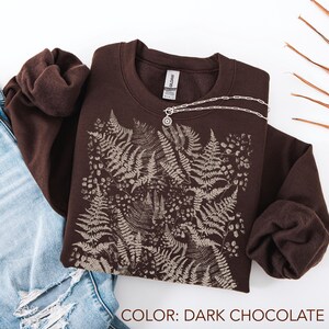 Puede incluir: Sudadera de color chocolate oscuro con un dise&ntilde;o de helechos y flores en un tono m&aacute;s claro. Un collar plateado con un colgante est&aacute; colocado encima. El texto "COLOR: DARK CHOCOLATE" est&aacute; en la parte inferior. La sudadera est&aacute; doblada.
