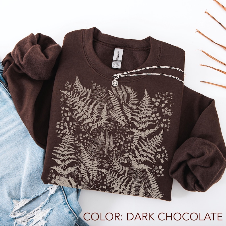 Puede incluir: Sudadera de color chocolate oscuro con un dise&ntilde;o de helechos y flores en un tono m&aacute;s claro. Un collar plateado con un colgante est&aacute; colocado encima. El texto "COLOR: DARK CHOCOLATE" est&aacute; en la parte inferior. La sudadera est&aacute; doblada.