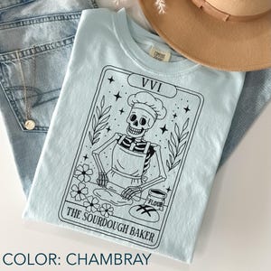 May include: A light blue t-shirt with a black graphic of a skeleton baker wearing a chef's hat and apron, rolling dough. The text "The Sourdough Baker" is below the image. The shirt is folded with a hat and jeans in the background.