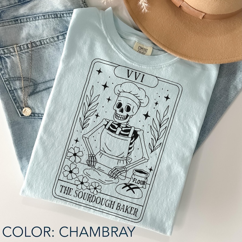 May include: A light blue t-shirt with a black graphic of a skeleton baker wearing a chef's hat and apron, rolling dough. The text "The Sourdough Baker" is below the image. The shirt is folded with a hat and jeans in the background.