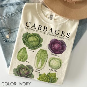 Puede incluir: Camiseta color marfil con una ilustración botánica de varios tipos de col, incluyendo col rizada y col roja. La camiseta muestra el texto "CABBAGES: A BOTANICAL STUDY OF BRASSICA VARIETIES."