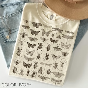 May include: Ivory t-shirt with a detailed print of insects, including butterflies, beetles, and dragonflies. The shirt has a crew neck and short sleeves. The text "COMFORT COLORS" is visible on the tag.