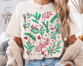 May include: Ivory-colored t-shirt featuring a festive botanical design. The design includes green holly leaves, red berries, and sprigs of red and green foliage. The shirt is a casual, short-sleeved style.
