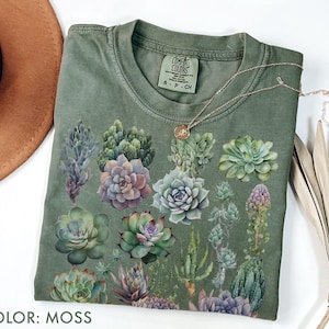 Op de afbeelding: Een mosgroen t-shirt met een aquarelontwerp van diverse vetplanten. Het shirt heeft een ronde hals en korte mouwen. Een gouden ketting met een kleine hanger is over het shirt gedrapeerd. De tekst "COLOR: MOSS" staat onderaan de afbeelding.